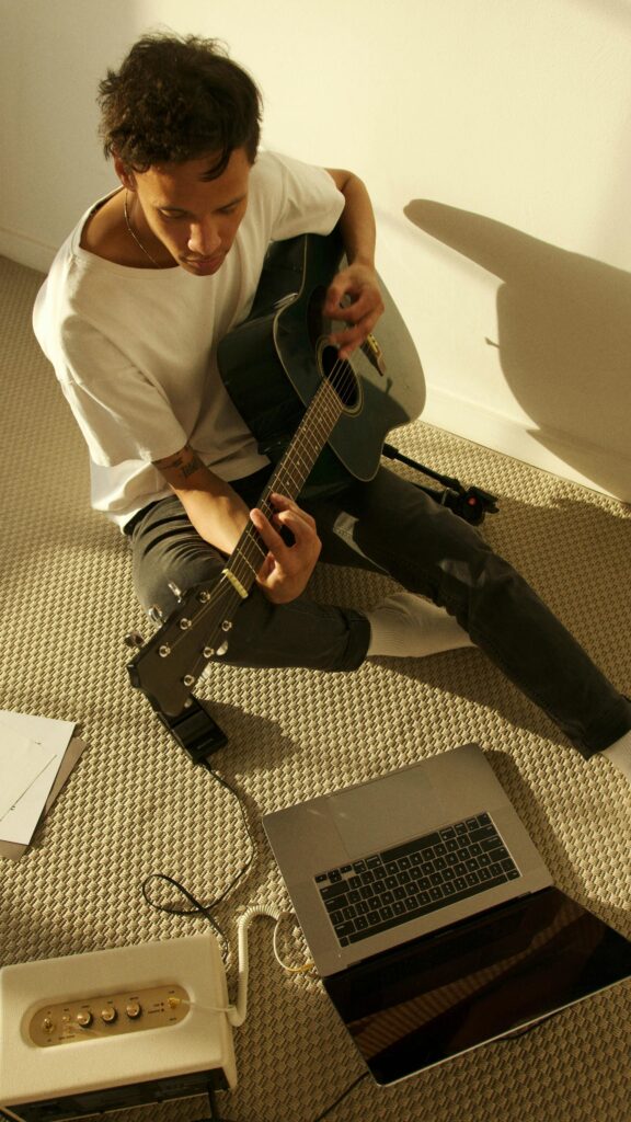 Young man composing music with an acoustic guitar and laptop indoors.